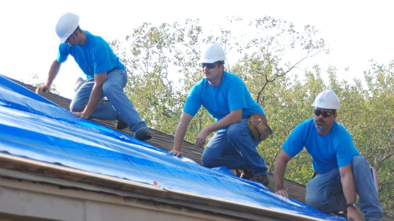 Three workers securing a blue tarp on a storm-damaged roof in Texas