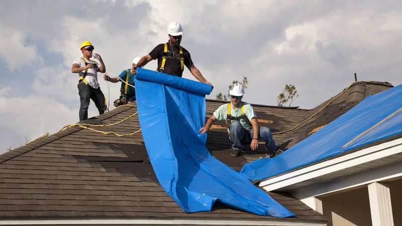 Roofers unrolling a blue tarp over a damaged roof