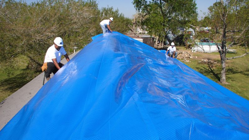 Workers fastening a blue tarp over a storm-damaged roof in Texas