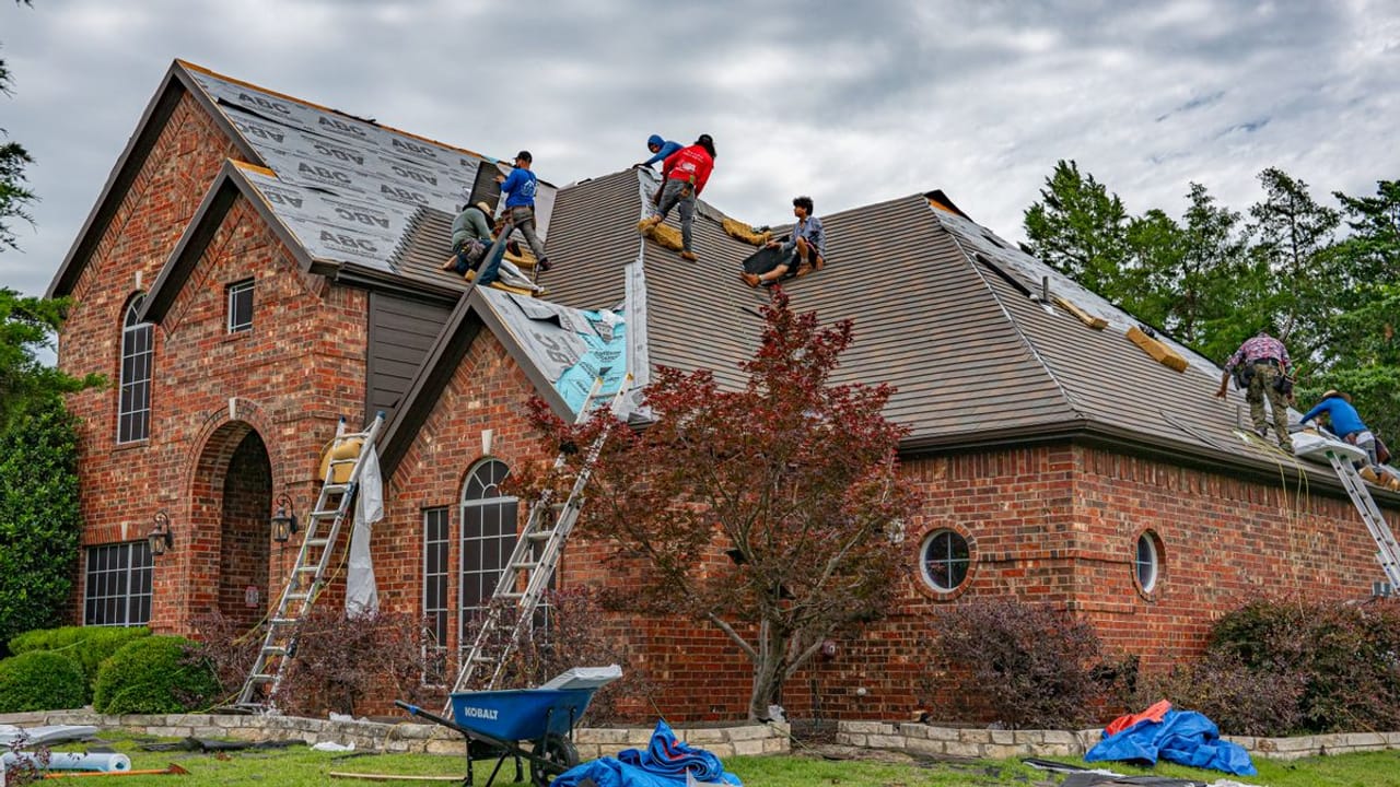 Storm Bolt Roofing crew replacing the asphalt shingle roof on a large brick Texas home, with ladders set and synthetic underlayment going down